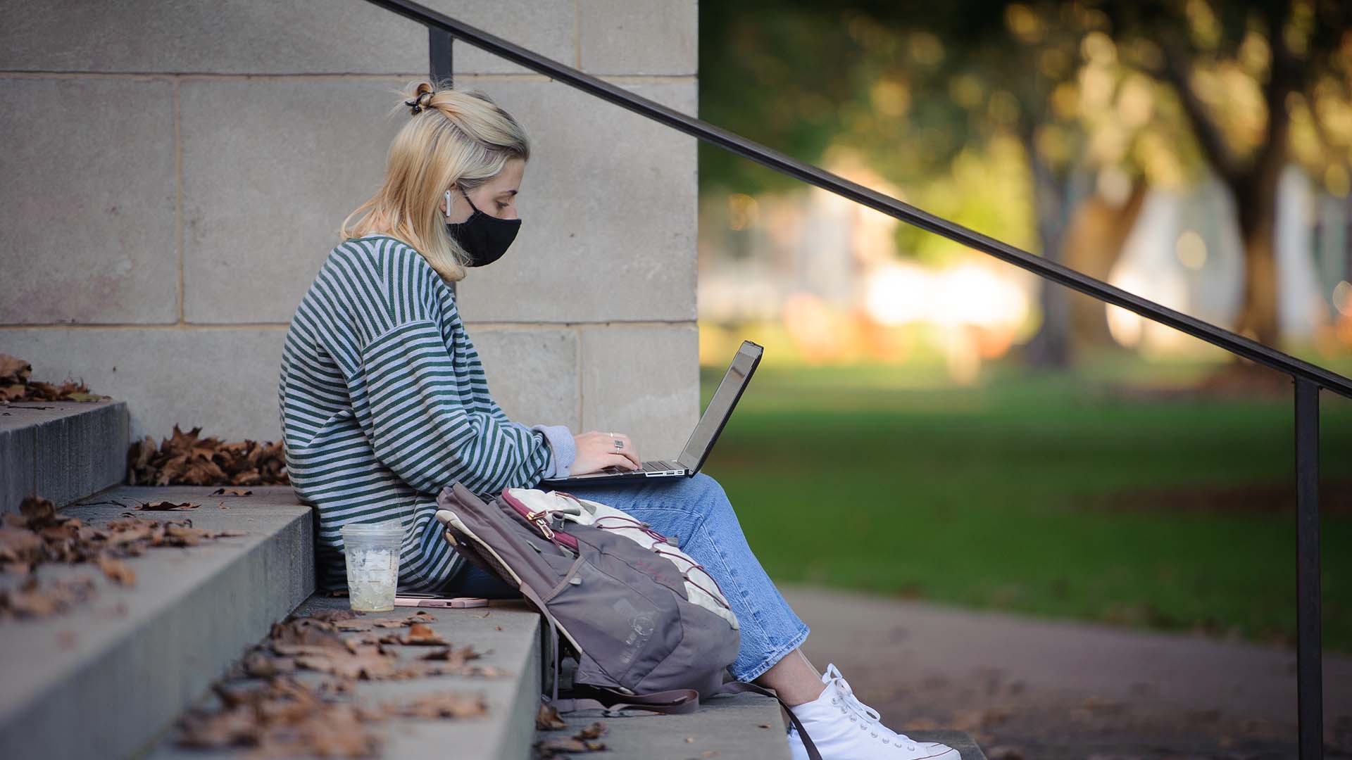 A student sitting outside on stairs with mask on looking at laptop
